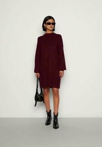 Vestido comprido de manga comprida em bordô, com gola alta, usado com botas pretas até ao tornozelo e óculos de sol, combinado com uma pequena mala preta.