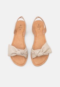 Sandales beige plates avec des sangles nouées sur les orteils et des sangles élastiques autour des talons, étiquetées "Call It Spring" à l'intérieur des semelles, marque végane.