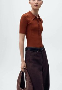Polo shirt marron côtelé à manches courtes avec une patte de boutonnage. Associé à un pantalon large marron foncé et un sac texturé avec des accents en métal.
