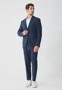 Σκούρο μπλε σκούρο ταγιέρ με μονόκουμπο σακάκι, γιακάδες με εγκοπές και στενά παντελόνια, σε συνδυασμό με λευκό πουκάμισο και λευκά αθλητικά παπούτσια.