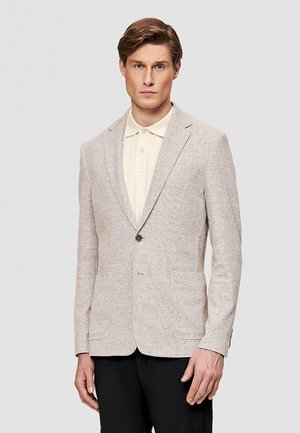 Mężczyzna w jasnobeżowej, teksturowanej marynarce, kremowej koszulce polo i czarnych spodniach, stojący na tle gładkiego, szarego tła.