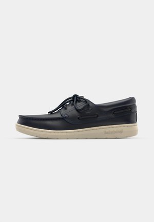 Zapato de barco de cuero azul marino con perfil bajo, suela de goma blanca, cordones redondos y costura decorativa en los laterales; cuenta con ojales de metal y logotipo.
