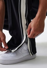 Pantalones deportivos negros con rayas verticales blancas y cremallera en el tobillo, combinados con unas zapatillas blancas. La tela tiene un acabado texturizado.