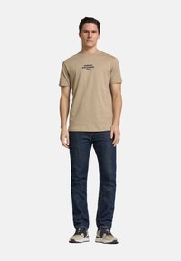 Beige bomulds t-shirt med sort tekstlogo, kombineret med mørkeblå jeans og lyse sportsko. Enkelt, tætsiddende design.