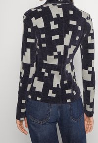 Kvinna som bär en åtsittande mörk kavaj med ett ljusgrått pixelmönster av kvadrater och blå denimjeans, sedd bakifrån.