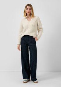 Flauschiger, kremfarbener Kabelstrickpullover mit V-Ausschnitt, kombiniert mit dunkelblauen, weit geschnittenen Jeans und silbernen Sneakers. Lässiges Outfit mit strukturierten Details.