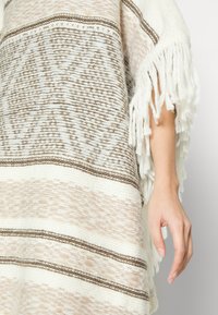 Poncho tecido em bege e creme com bordas de franjas. Apresenta padrões geométricos e riscas texturizadas em materiais suaves e quentes.