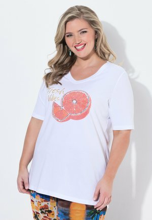 Hymyilevä vaaleatukkainen nainen, jolla on laineikkaat hiukset, pukeutunut valkoiseen paitaan, jossa lukee ”Fresh Vibes”, ja oranssin viipaleita, värikkäät kuvioidut housut, seisoo valkoista seinää vasten.