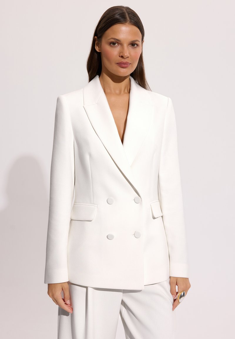 Kobieta ubraną w biały, dwurzędowy blezer z dopasowanymi białymi spodniami, stojąca na jednolitym, jasnym tle.