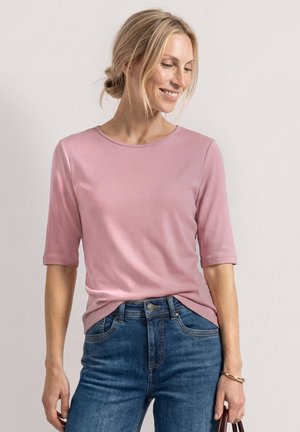 Kvinde med blond hår i en lav knold bærer en lyserød bluse med albuelange ærmer og blå jeans, smiler og kigger nedad til siden.