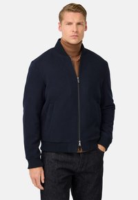 Moški v temno modri zadrgavi jakni nad rjavim turtleneckom in temnimi kavbojkami, stoji z eno roko v žepu ob enobarvni ozadju.