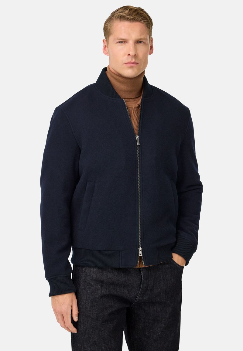 Moški v temno modri zadrgavi jakni nad rjavim turtleneckom in temnimi kavbojkami, stoji z eno roko v žepu ob enobarvni ozadju.