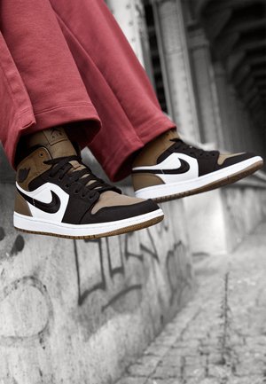 Jambes portant un pantalon rose et des baskets Nike Air Jordan 1 marron, noir et blanc suspendues au-dessus d'un rebord en béton couvert de graffiti.