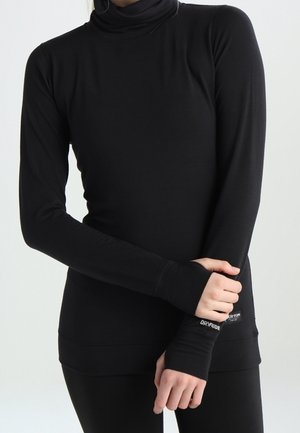 Persona que lleva una camiseta ajustada negra de manga larga con cuello alto y agujeros para los pulgares, sujetando una muñeca frente a su cuerpo.