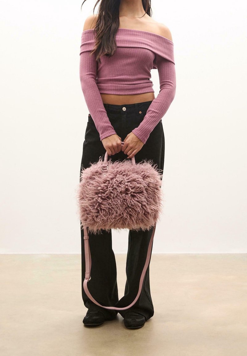 Femme portant un top rose côtelé à épaules dénudées et un pantalon noir, tenant un sac à main rose duveteux avec des poignées et une longue bandoulière.