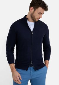 Navy blaues Zip-Cardigan aus Strickstoff, mit Stehkragen, langen Ärmeln und zwei Vordertaschen. Getragen mit einem weißen T-Shirt und blauen Hosen.