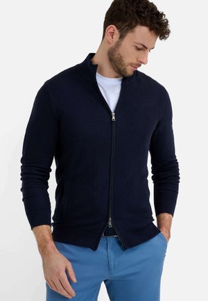 Navy blaues Zip-Cardigan aus Strickstoff, mit Stehkragen, langen Ärmeln und zwei Vordertaschen. Getragen mit einem weißen T-Shirt und blauen Hosen.
