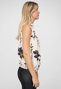 Mouwloze blouse in crèmekleurige stof met zwarte bloempatronen. Heeft een gerimpelde zoom. Gepaard met zwarte strakke leggings.