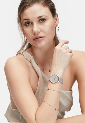 Helle graue Uhr mit rundem Zifferblatt, silbernen Akzenten und Silikonarmband. Modell trägt zarte roségoldene Armreifen und ein strukturiertes beiges Oberteil.