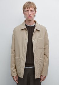Giacca beige con zip e bottoni a pressione, dotata di tasca frontale e due tasche laterali, indossata sopra un maglione a coste scuro e una camicia grigia.