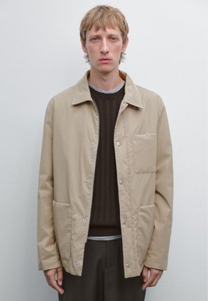 Beige Zip-Jacke mit Druckknöpfen, ausgestattet mit einer Fronttasche und zwei Seitentaschen, getragen über einem dunklen Rippenpullover und einem grauen Hemd.
