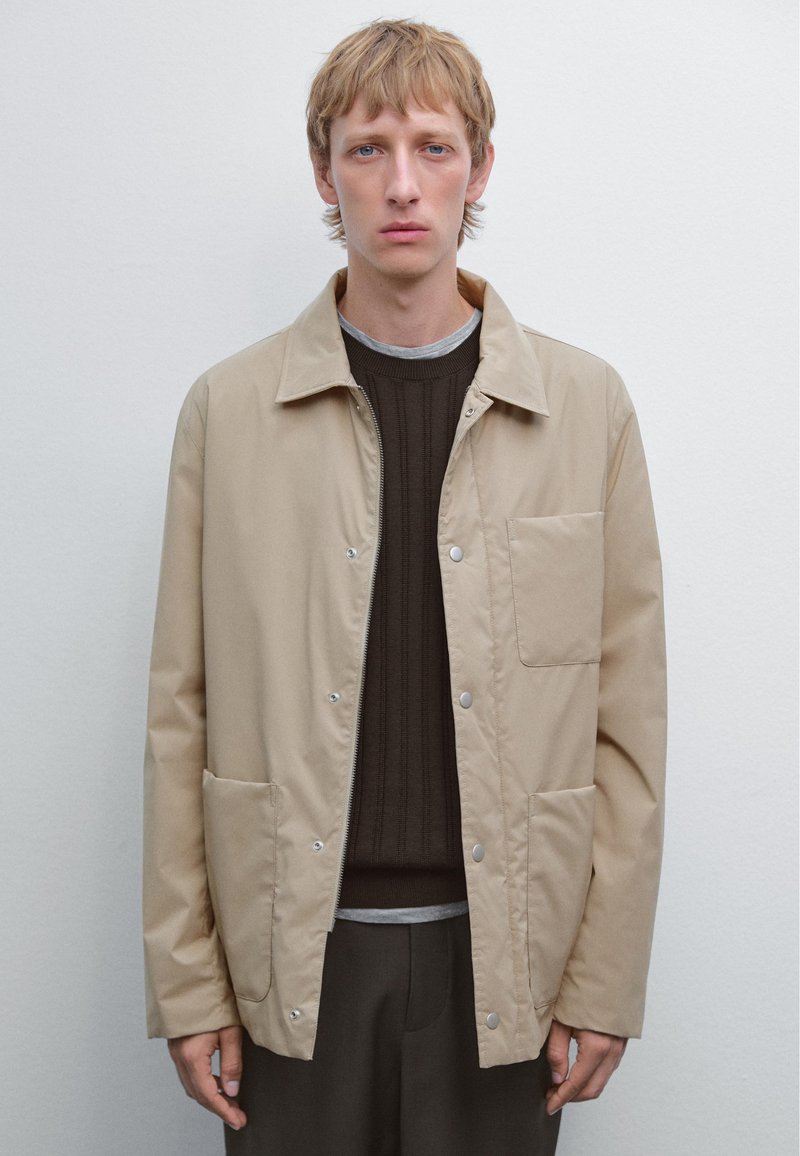 Beige ritsjack met drukknoopjes, met een voorzak en twee zijzakken, gedragen over een donker geribbeld sweater en een grijze shirt.