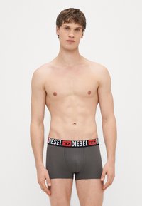 Pánske boxerky v sivej látke, s čiernym pásikom so sivým červeným logom "DIESEL". Dizajn je priliehavý s hladkou textúrou.
