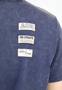 Temno modra majica s teksturirano tkanino, ki vsebuje tri prišite oznake: "ADR SPRING 2025," "RE-CREATE," in "Atelier de Roupa, ustanovljen 2018."
