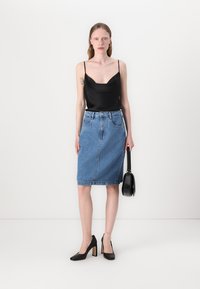 Top in satin nero con scollatura profonda, abbinato a una gonna a tubo in denim blu chiaro. La modella tiene una piccola borsa nera e indossa tacchi neri.