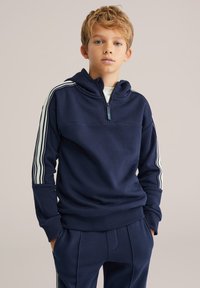 Tumši zils puslokveida džemperis ar baltām svītrām gar piedurknēm, ribošiem manžetēm un vaļēju piegriezumu. Mīksta tekstūra, daļa no saskaņota komplekta.