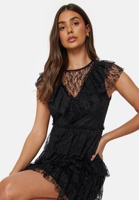 Robe noire en dentelle avec des détails froncés, des manches courtes et un décolleté en V. La texture est composée d'effets de tissus superposés, transparents et opaques.