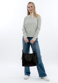 Vrouw in gestreept shirt met lange mouwen en spijkerbroek die met beide handen een grote zwarte handtas vasthoudt voor een witte achtergrond.