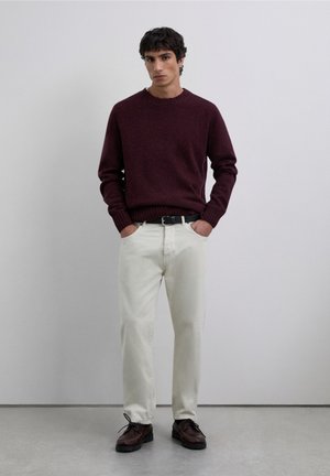 Maglione bordeaux a maglia con collo a coste, pantaloni di cotone color crema e scarpe marroni stringate con suola texturizzata. Sfondo neutro.