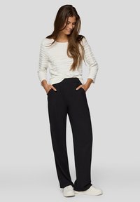 Frau trägt ein strukturiertes weißes Langarmshirt, schwarze weit geschnittene Hose, weiße Sneakers, steht mit Händen in den Taschen, lächelt und schaut nach unten.