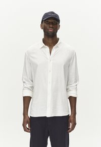 Weißes Leinen-Hemd mit Knopfverschluss und hochgerollten Ärmeln, kombiniert mit einer dunkelblauen Kappe mit Logo. Das Modell steht vor einem schlichten weißen Hintergrund.