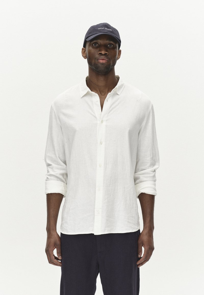 Weißes Leinen-Hemd mit Knopfverschluss und hochgerollten Ärmeln, kombiniert mit einer dunkelblauen Kappe mit Logo. Das Modell steht vor einem schlichten weißen Hintergrund.