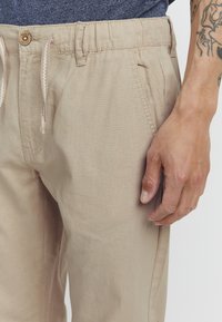 Beige elastische Bundhosen mit einem Kordelzug, seitlichen Taschen und einer glatten Textur. Mit einem Knopfverschluss und kontrastierenden Nähten.