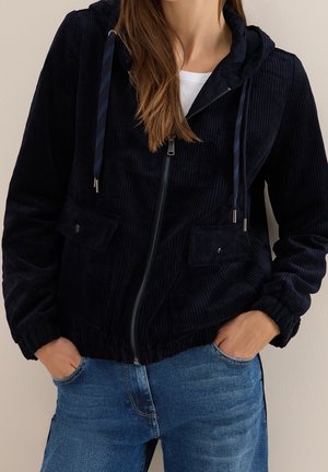 Femme portant un sweat à capuche zippé en velours côtelé bleu marine avec cordons et un jean bleu, les mains dans les poches, sur un fond uni.