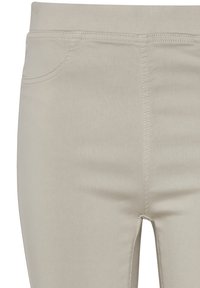 Gros plan sur un pantalon beige montrant la couture avant, la ceinture et une partie de la poche avant sur un fond uni.