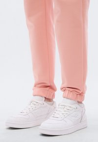 Ténis brancos com detalhes texturizados, combinados com calças de jogging cor de rosa claro com punhos elásticos. O design é desportivo e casual.