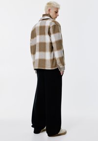 Person med kort blont hår som bär en brun och vit rutablickjacka, lösa svarta byxor och beige sneakers, står framför en vit bakgrund.