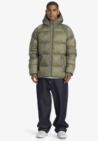 Olivgrön puffjacka med huva, dragkedja fram och två sidofickor; tillverkad av glänsande, strukturerat material; kombinerad med mörka jeans.