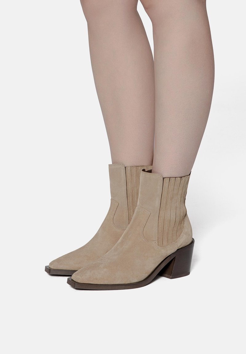 Beige nubukové kotníkové boty s špičatou špičkou, elastickými bočními panely a silnými tmavohnědými blokovými podpatky nošené na holých nohou.