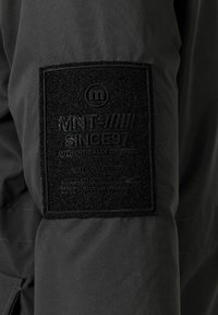 Svart jacka med en matt svart tygpatch på ärmen, präglad med "MNT SINCE 97" och en cirkulär logotyp. Texturerad yta synlig.