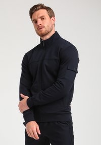 Veste zippée bleue marine en tissu doux, dotée d'un col haut, de poches avant et d'un badge sur la manche gauche. Texture lisse.