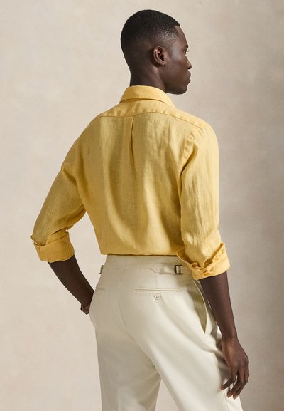 Κίτρινο λινό πουκάμισο με σηκωμένα μανίκια και άνετη εφαρμογή, σε συνδυασμό με ανοιχτό κρεμ παντελόνι με ραμμένες τσέπες στο πίσω μέρος και ζώνη.