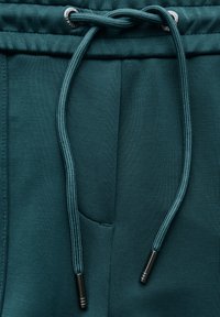 Teal joggers med dragsko i midjan och metalldelar, samt en sidoficka. Mjuk textur med en subtil lyster.