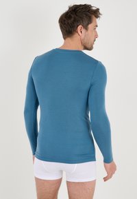 Camicia a maniche lunghe, aderente, di colore teal, realizzata in tessuto liscio. Presenta un colletto a girocollo e polsini a costine, abbinata a slip bianchi.