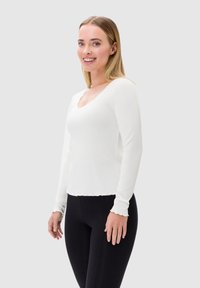 Biała, długorękawowa bluzka z prążkowanego materiału, z falbaniastym dekoltem i mankietami, o dopasowanym kroju. W połączeniu z czarnymi legginsami.
