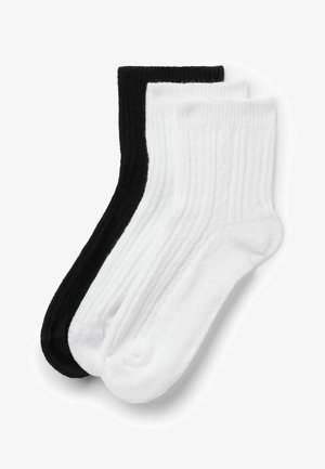 Due paia di calzini a costine, uno nero e uno bianco, distesi e sovrapposti su uno sfondo bianco.